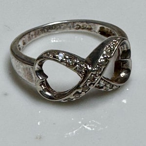 May include: A silver-toned ring featuring an infinity symbol design. The ring is adorned with small, clear gemstones along the curves of the infinity symbol. The ring is set on a white surface.