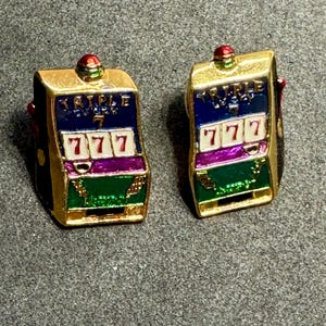 May include: A pair of gold-toned slot machine earrings. Each earring features a colorful design with the text "TRIPLE 7" above three "777" symbols. The earrings have red, green, purple, and blue accents, with a small red gem on top.