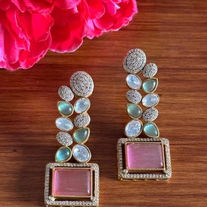 May include: A pair of gold earrings featuring pink and green stones. The earrings are made of metal and have a square design. The stones are set in a pave setting.