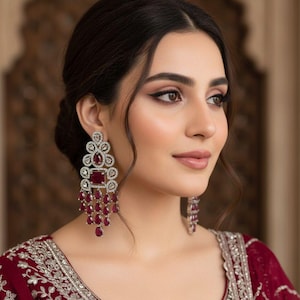 Peut inclure: Boucles d'oreilles pendantes élégantes avec un sertissage argenté et des pierres précieuses rouge rubis. Les boucles d'oreilles présentent un groupe de petits bijoux suspendus sous une pierre rectangulaire plus grande, avec un design en filigrane détaillé. La modèle porte une robe bordeaux avec broderie argentée.