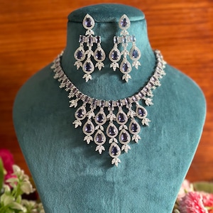 May include: A silver necklace and earrings set with a teardrop design. The necklace features a cascade of pear-shaped purple stones surrounded by smaller clear stones. The earrings are similar in design, with a dangling teardrop purple stone.
