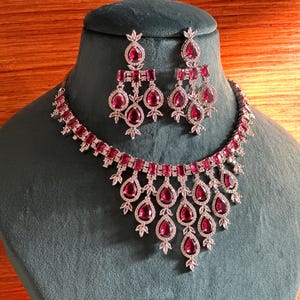 May include: A silver necklace and earrings set with a teardrop design. The set features large, red, pear-shaped stones surrounded by smaller, clear stones.