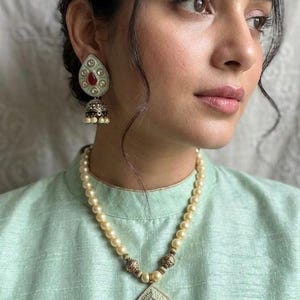May include: A pearl necklace and matching earrings with a teardrop pendant and earring design. The pendant and earrings feature a red gemstone centre, surrounded by a white and gold design. The jewellery is worn with a light green top.