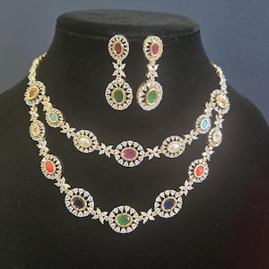May include: A gold-toned necklace and earring set. The necklace features two strands with colorful oval gemstones and clear accent stones. The earrings have a matching design. The jewelry is displayed on a black mannequin.