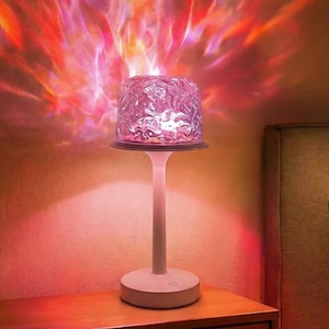 May include: A white table lamp with a textured, pink lampshade. The lamp projects a colorful light pattern onto the wall, creating a warm, ambient glow. The light display includes shades of red, orange, and pink.