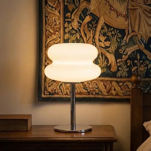 May include: A white, retro-style table lamp with a stacked, rounded shade. The lamp has a chrome base and stem, and sits on a wooden nightstand. The background features a tapestry and a bed.