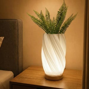 May include: A white, swirled ceramic vase with a wooden base, illuminated from within, holding green fern-like plants. The vase sits on a wooden nightstand next to a bed with a gray headboard. The lamp provides soft, ambient lighting.
