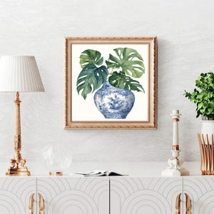 Puede incluir: Una pintura de acuarela de un jarr&oacute;n de porcelana azul y blanco con hojas verdes de monstera. El jarr&oacute;n est&aacute; decorado con un patr&oacute;n floral. La pintura est&aacute; enmarcada en un marco dorado.