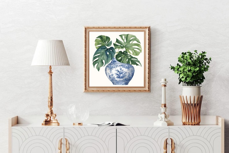 Puede incluir: Una pintura de acuarela de un jarr&oacute;n de porcelana azul y blanco con hojas verdes de monstera. El jarr&oacute;n est&aacute; decorado con un patr&oacute;n floral. La pintura est&aacute; enmarcada en un marco dorado.