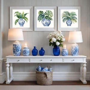 Puede incluir: Tres impresiones enmarcadas de pinturas de acuarela de plantas tropicales en jarrones de porcelana azul y blanco. Las impresiones se muestran en una pared blanca sobre una consola blanca con l&aacute;mparas, jarrones y tarros de porcelana azul y blanco.