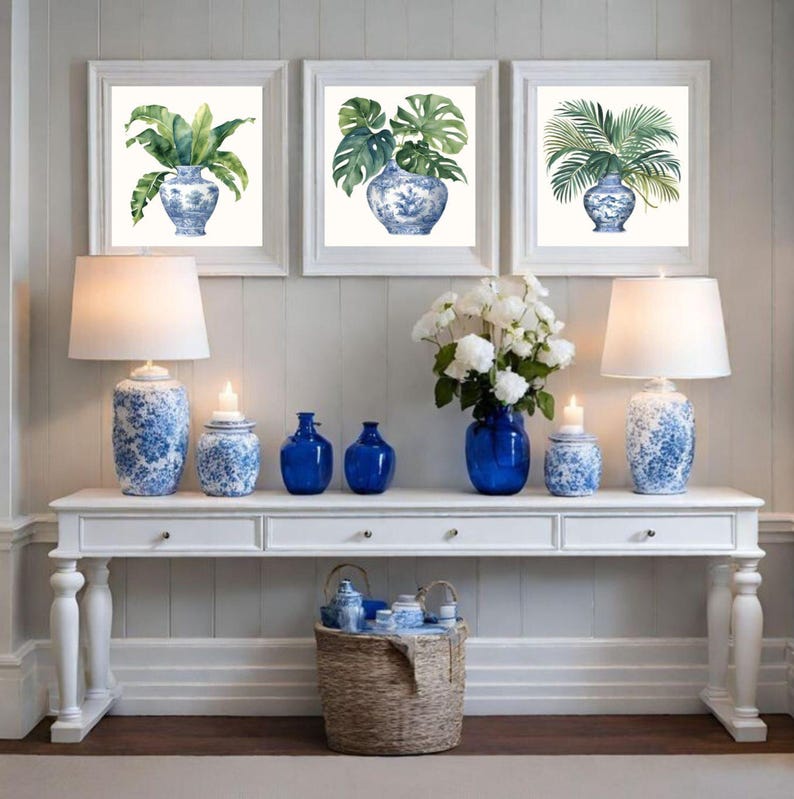 Puede incluir: Tres impresiones enmarcadas de pinturas de acuarela de plantas tropicales en jarrones de porcelana azul y blanco. Las impresiones se muestran en una pared blanca sobre una consola blanca con l&aacute;mparas, jarrones y tarros de porcelana azul y blanco.