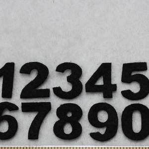 May include: Black felt numbers 1 through 0 arranged on a light gray surface. The numbers are in a bold, sans-serif font. A ruler is visible at the bottom of the image, providing a size reference.