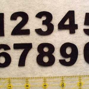 May include: Black felt numbers 1 through 0, cut out and arranged in two rows. The numbers are on a white surface with a yellow ruler underneath.