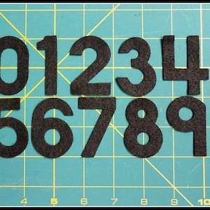 May include: A collection of black felt numbers from zero to nine, arranged on a green grid background. The numbers are in a bold, sans-serif font, and each is distinct and easily readable. The felt material gives them a soft, textured appearance.
