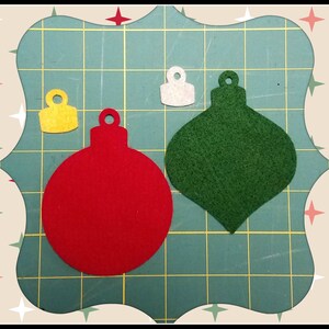 May include: A collection of felt Christmas ornaments in red, green, and yellow. The ornaments are in various shapes, including a round ball and a teardrop shape. The background is a green cutting mat with a decorative border.