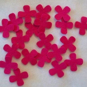 May include: A collection of 18 bright pink felt flower shapes, each with five petals. The flowers are scattered on a white surface.