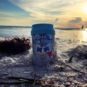 May include: A can of Pabst Blue Ribbon beer with a blue lid, partially submerged in the sand on a beach. The can is surrounded by seaweed and shells. The ocean is in the background, with a bright sun shining in the sky.