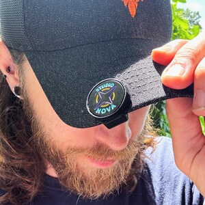 May include: Black trucker hat with an orange phoenix logo and a Studio Nova phone grip. The phone grip is black with a circular design featuring the words "Studio Nova" and a starburst pattern in blue and yellow.