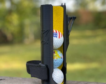 May include: A black golf ball holder with a yellow interior and a strap. Three golf balls are inside the holder, one with an American flag design, one with a world map design, and one with a white and black speckled design.