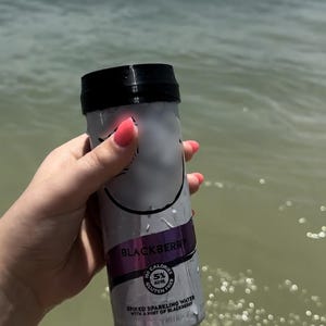 May include: A silver can of blackberry flavored spiked sparkling water with a black lid. The can has the text "Blackberry" and "Spiked Sparkling Water with a hint of Blackberry" printed on it. The can is being held by a hand in the water.