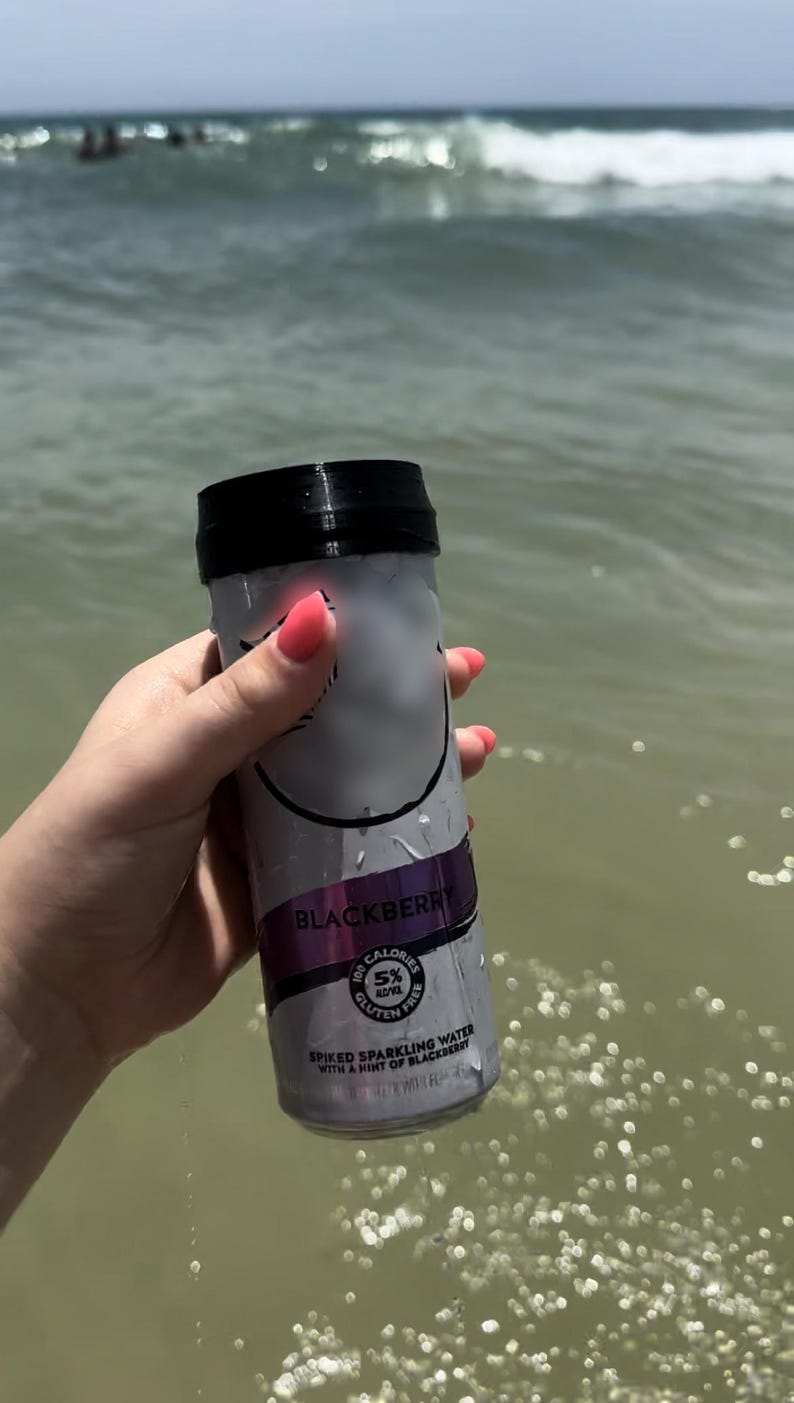 May include: A silver can of blackberry flavored spiked sparkling water with a black lid. The can has the text "Blackberry" and "Spiked Sparkling Water with a hint of Blackberry" printed on it. The can is being held by a hand in the water.