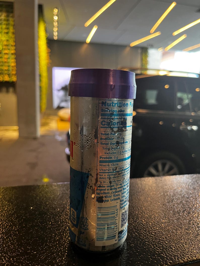 May include: A silver can of energy drink with a purple top and a nutrition facts label. The can is wet and has condensation on it. The can is sitting on a black surface.