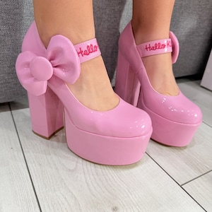 Pode incluir: Um par de sapatos de plataforma de verniz rosa claro com salto bloco grosso, biqueira arredondada e um laço grande a condizer na lateral. A tira do tornozelo inclui o texto Hello Kitty bordado com fio rosa na tira frontal.