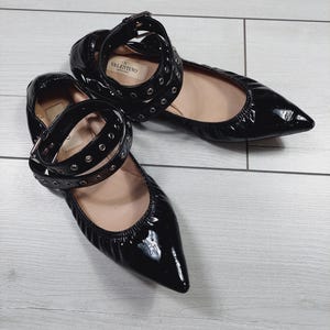 Może przedstawiać: Para czarnych baletek ze sk&oacute;ry lakierowanej ze spiczastymi noskami. Buty posiadają paski na kostkę ze srebrnymi oczkami i klamrami. Logo Valentino jest widoczne na pasku. Buty leżą na jasnej powierzchni imitującej drewno.