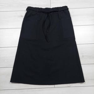 Puede incluir: Falda negra con dos bolsillos delanteros y cintura con cordón. La falda está hecha de una tela negra lisa y tiene un dobladillo a la altura de la rodilla. La falda se muestra sobre una superficie clara de vetas de madera.