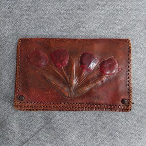 Puede incluir: Una cartera de cuero marrón con un diseño floral. La cartera presenta un patrón en relieve de cuatro tulipanes en varios tonos de rojo. Los bordes están cosidos con un patrón decorativo y asegurados con dos botones marrones.