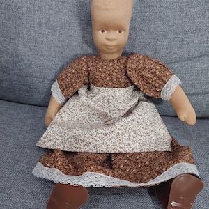 Puede incluir: Muñeca hecha a mano con cabeza y cuerpo de tela, vestida con un vestido marrón con estampado floral y ribete de encaje blanco. La muñeca tiene zapatos de plástico marrones y está sentada sobre una superficie gris.