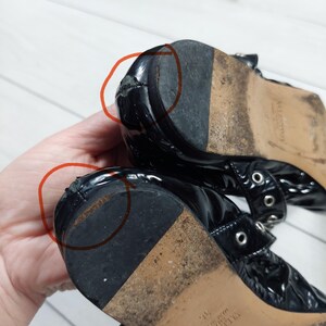 Może przedstawiać: Zbliżenie dw&oacute;ch czarnych lakierowanych but&oacute;w, pokazujące zużycie na obcasach. Buty mają zapięcie na klamrę i prawdopodobnie są designerskie. Podeszwy są zużyte, co wskazuje na wcześniejsze użytkowanie.