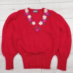 Puede incluir: Un suéter rojo con cuello en V y diseño floral alrededor del cuello. Las flores son blancas, rosas y verdes. El suéter tiene mangas largas con puños ajustados y un dobladillo acanalado.