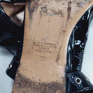 Może przedstawiać: Zbliżenie na podeszwę czarnego buta ze sk&oacute;ry lakierowanej. Podeszwa jest zużyta i pokazuje logo Valentino Garavani, "Made in Italy" oraz rozmiar 36. But ma paski z metalowymi oczkami.