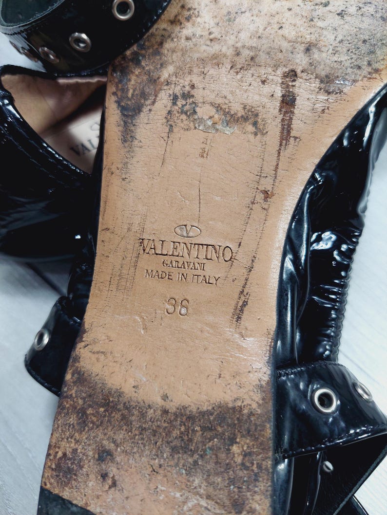 Może przedstawiać: Zbliżenie na podeszwę czarnego buta ze sk&oacute;ry lakierowanej. Podeszwa jest zużyta i pokazuje logo Valentino Garavani, "Made in Italy" oraz rozmiar 36. But ma paski z metalowymi oczkami.