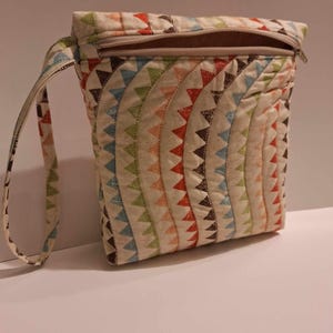 May include: A quilted pouch with a colorful geometric pattern. The pouch features a zipper closure and a small handle. The design includes rows of triangles in shades of red, orange, green, blue, and brown on a cream background.