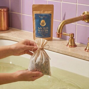 Peut inclure: Un sachet de thé de bain en mousseline beige, noué avec de la ficelle, est tenu au-dessus d'une baignoire. Un sac en papier brun avec le texte "SACRED WATERS" et "Menstrual Comfort Bath Tea" est en arrière-plan. Le robinet est doré.