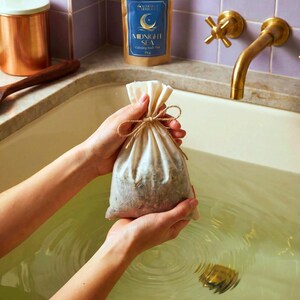 Peut inclure: Un sac de thé de bain en mousseline beige, noué avec de la ficelle, est tenu au-dessus d'une baignoire remplie d'eau. Un paquet en papier brun avec une étiquette bleue indiquant "Midnight Sea Calming Bath Tea" est en arrière-plan. Un récipient en cuivre et un robinet sont également visibles.
