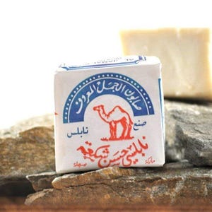 Puede incluir: Una pastilla de jabón blanco con una etiqueta azul y roja que muestra un camello y texto árabe. El texto de la etiqueta dice "صابون الجمل" que se traduce como "Jabón de camello".