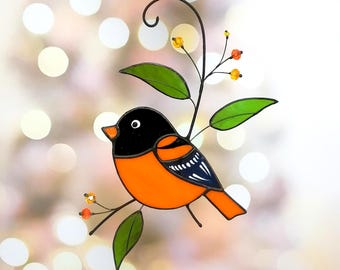 Puede incluir: Un adorno de vidrio tintado que representa un pájaro naranja brillante con marcas negras y azules posado en una rama verde con bayas naranjas. El pájaro tiene la cabeza negra y un ojo blanco.