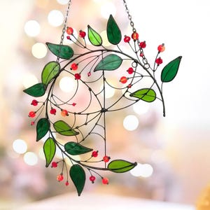 May include: A stained glass suncatcher featuring a crescent moon shape with a spiderweb design. The moon is adorned with green leaves and red beads.