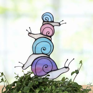 May include: A stained glass art piece featuring four stacked snails with translucent shells in shades of light blue, pink, medium blue, and purple. Each snail has a white body with black wire outlines and antennae, creating a vertical tower.