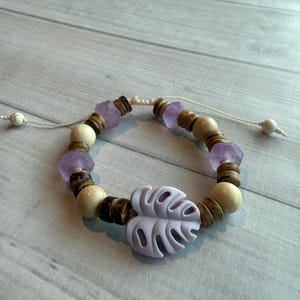 May include: A bracelet featuring a light purple leaf-shaped charm and a variety of beads. The beads include light wood, dark brown, and faceted purple stones. The bracelet is strung on a white cord and has adjustable ends.