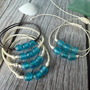 May include: Several handmade bracelets with blue glass beads and off-white cords are displayed on a wooden surface. A seashell and sea glass pieces are also present, suggesting a beach or nautical theme.