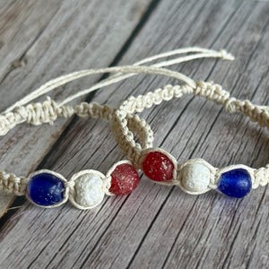 May include: Two macrame bracelets crafted from light beige cord, each featuring three round beads in red, white, and blue. The bracelets have adjustable cord ties and are arranged in a circular pattern, showcasing a knotted texture throughout.