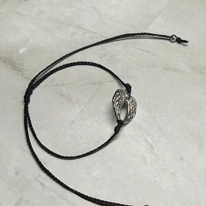 May include: A silver-coloured pendant shaped like angel wings hangs from a black cord necklace. The cord features adjustable sliding knots and small silver beads at the ends. The necklace is shown on a light-coloured surface.