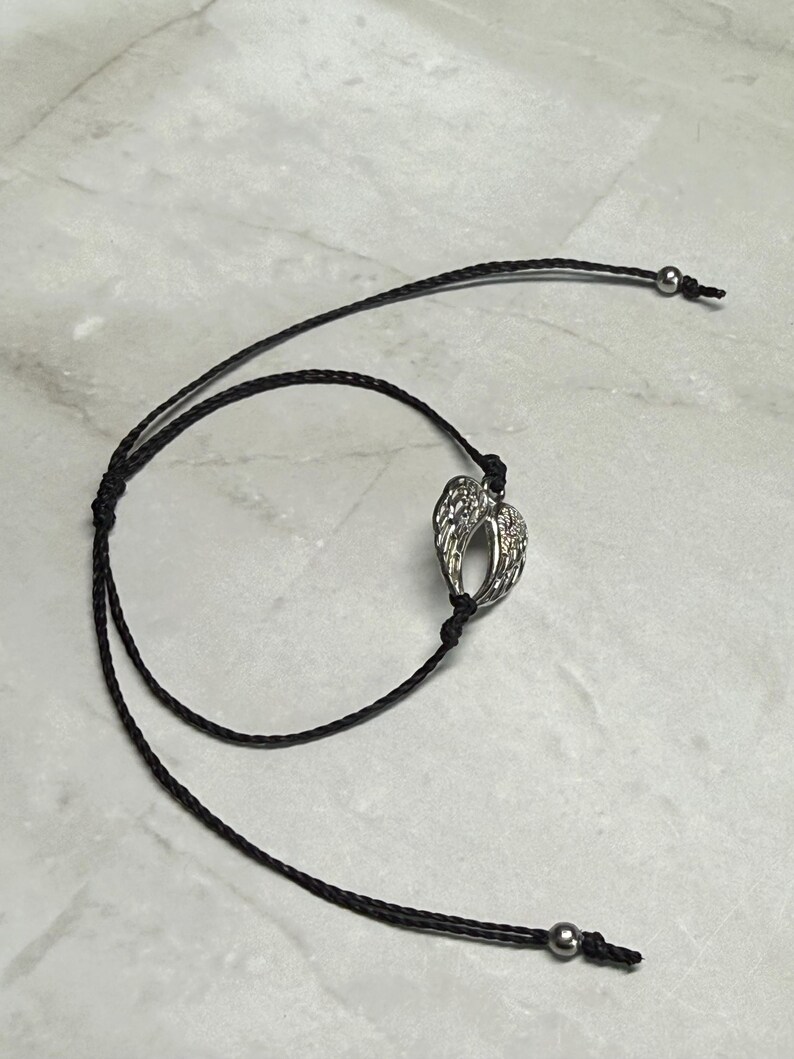 May include: A silver-coloured pendant shaped like angel wings hangs from a black cord necklace. The cord features adjustable sliding knots and small silver beads at the ends. The necklace is shown on a light-coloured surface.