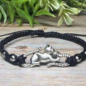 May include: Black macrame bracelet featuring a silver cat charm. The bracelet has a woven design with silver beads. The cat charm is detailed and adds a unique touch to the accessory. The bracelet is adjustable.