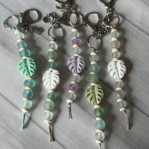 May include: Five beaded keychains with silver-tone clasps. Each keychain features a strand of beads in shades of green, white, and purple, with a decorative leaf charm. The keychains have a tropical theme.