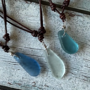 May include: Three necklaces featuring smooth, frosted sea glass pendants in shades of deep blue, pale white, and teal. Each pendant is attached to a dark brown leather cord with a silver-toned wire wrap and adjustable sliding knots for length.
